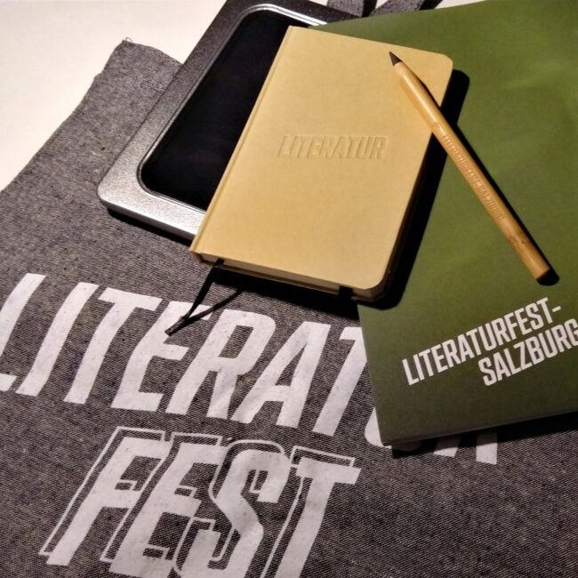 Stoffsackerl mit Aufdruck "Literaturfest Salzburg", auf dem ein Notizbuch mit Aufdruck "Literatur" und ein Stift mit Aufdruck "Literaturfest Salzburg", Metallbox und Programmheft liegen