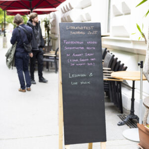 Schild vor dem Toihaus Theater, auf dem steht "Literaturfest Salzburg 2025. Festzentrum wegen Schlechtwetter heute im Academy Salon (1. Stock, Eingang über die Academy-Bar). Lesungen & Live Musik"